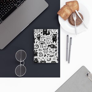 May include: A notebook with a black and white monster doodle pattern is displayed on a desk next to a laptop, glasses, and a plate of food. The notebook's cover features various cartoon monsters and creatures.