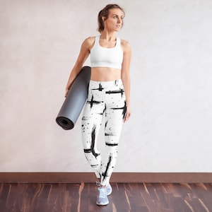 Może przedstawiać: Biały biustonosz sportowy i legginsy z czarnym wzorem pociągnięć pędzla. Legginsy mają wysoki stan i wzór czarnych pociągnięć pędzla i rozprysków. Zwinięta szara mata do jogi jest trzymana.