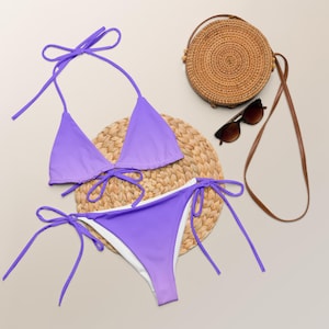 May include: A lavender bikini set with tie closures, a matching round woven bag, and sunglasses. The bikini top is a triangle style, and the bottoms have white lining. The bag has a brown strap.