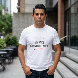 May include: White t-shirt with the text "my status report says green. MY FACE SAYS OTHERWISE." in black font. The text is centered on the shirt. A red line underlines the word "OTHERWISE".