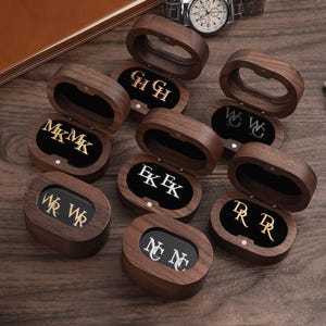 May include: A collection of personalized cufflinks in wooden boxes, each displaying initials in gold or silver against a black velvet interior. The open boxes are arranged on a dark wooden surface, with a watch and decorative elements in the background.