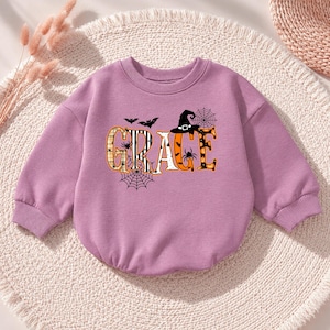 Pode incluir: Uma camisola de manga comprida cor de lavanda com o nome GRACE em letras grandes e decorativas. As letras estão preenchidas com padrões de xadrez, riscas e abóboras cor de laranja, acentuadas com teias de aranha, aranhas, morcegos e um chapéu de bruxa pretos.