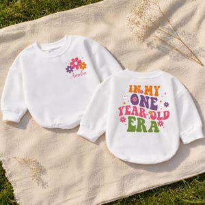 May include: Two white long-sleeve sweatshirts. One features a small floral graphic with the name Amelia. The other displays the text IN MY ONE YEAR OLD ERA in colorful, bold lettering surrounded by small flower icons and star shapes.