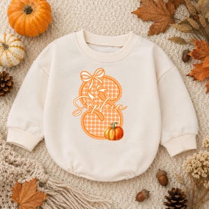 May include: A cream-colored long-sleeve sweatshirt featuring a large orange and white checkered letter S with a bow on top. The name Sophia is written in cursive across the center of the letter, and a small orange pumpkin sits at the bottom.