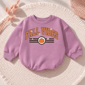 Pode incluir: Uma camisola de manga comprida em tom malva com uma impressão gráfica com o texto FALL VIBES em letras laranja, uma ilustração central de uma abóbora laranja e pequenas margaridas brancas. Gola redonda e punhos canelados.