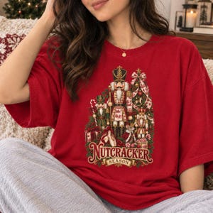 May include: A red short-sleeved t-shirt featuring a graphic print of three nutcracker figures, a decorated Christmas tree, and a mouse wearing a crown. The text "NUTCRACKER SEASON" is printed in gold and red lettering at the bottom of the design.