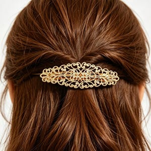 May include: A gold-colored filigree hair clip is secured in the back of brown hair. The clip has an ornate, symmetrical design with floral and scroll details. The hair accessory is a barrette style.