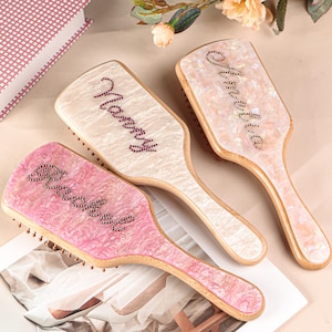 Könnte beinhalten: Drei personalisierte Haarbürsten mit Holzgriffen und dekorativen Oberflächen. Eine ist rosa mit dem Namen "Brooke" in Strasssteinen, eine andere ist cremefarben mit "Nancy" und die dritte ist cremefarben mit "Amelia".