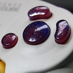 May include: Close-up of a white GameCube controller with custom buttons. The buttons are a gradient of purple and blue with a starry effect. The controller has a yellow joystick.