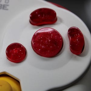 May include: Close-up of a white Nintendo GameCube controller with red buttons. The controller has a start/pause button and a yellow joystick. The red buttons are translucent.