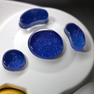 May include: Close-up of a white game controller with four blue, glittery buttons. The buttons are in various shapes, including a circle, a bean shape, and a rounded rectangle. The controller has the text "NINTENDO CUBE" printed on it.