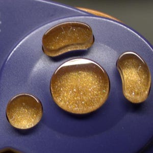 May include: Close-up of a blue video game controller with four gold-glittered buttons. The buttons are various shapes, including round, oval, and a kidney bean shape. The word "CUBE" is visible on the controller.