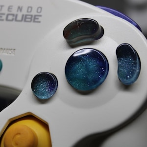 May include: Close-up of a white Nintendo GameCube controller with custom buttons. The buttons are a mix of blue, teal, and purple, with a galaxy-like design. The text "NINTENDO GAME CUBE" and "START/PAUSE" are visible.