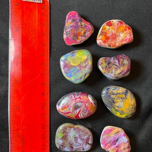 May include: A collection of seven painted rocks in various shapes and sizes. The rocks feature vibrant colors like pink, yellow, blue, and orange, with swirling patterns. A red ruler, marked in inches, is positioned next to the rocks.