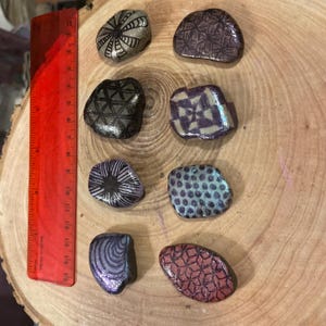 May include: A collection of seven decorative, hand-painted stones with various geometric patterns in black, silver, purple, and teal. A red ruler with inch markings is visible on the left side, indicating size.