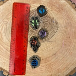 May include: Five hand-painted decorative stones with nature-inspired designs. The stones feature colorful illustrations of leaves and plants. A red ruler, marked in inches and centimeters, is placed next to the stones. The stones are on a wooden surface.