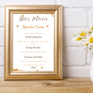 May include: A framed bar menu with a gold-colored frame. The menu features the text "Bar Menu" and "Signature Drinks" in elegant script. Drink names include "The Maple Whiskey Sour" and "The Cozy Cider Mule". The background is white.