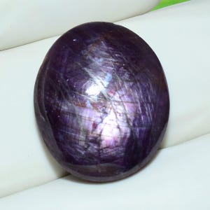 Op de afbeelding: Een gepolijste, ovaalvormige cabochon edelsteen met een diepe, donkerpaarse tint met zichtbare interne insluitingen en een subtiele, lichtreflecterende glans over het gladde, afgeronde oppervlak, met natuurlijke variaties in kleur en textuur.