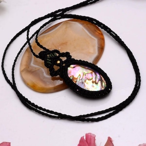 May include: A black macrame necklace featuring an oval pendant with an iridescent abalone shell. The shell displays a mix of colors, including pink, green, and gold. The necklace is displayed on a light brown stone.