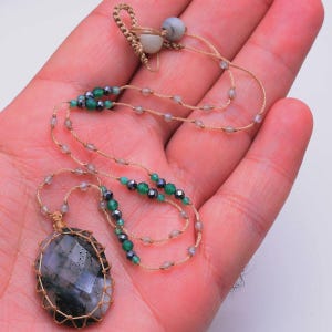 Op de afbeelding: Een handgemaakte ketting met een grote, facetgeslepen, ovale hanger in een geweven, beige koord. De ketting heeft een beige koord met kleine, heldere kralen en groene en zwarte kralen. Een uniek sieraad.