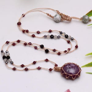Può includere: Una collana con un grande ciondolo in pietra rotonda viola scuro racchiuso in un cordino di macramè color marrone chiaro. Il cordino è decorato con piccole perline sfaccettate rosso granato, perline grigie traslucide e perline grigio metallizzato.
