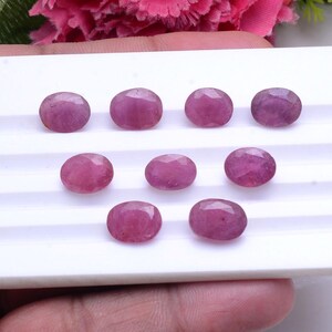 Può includere: Una collezione di nove rubini rosa sfaccettati di forma ovale, disposti in tre file su un vassoio espositore bianco. Ogni gemma presenta una tonalità rosa intenso traslucida con una superficie lucida che riflette la luce.