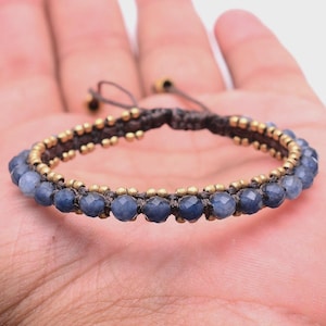 May include: A handmade bracelet featuring dark blue faceted beads and small gold-colored beads. The bracelet is strung on a dark brown cord and has an adjustable closure. The bracelet is shown on a person's hand.