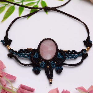 Op de afbeelding: Een macramé ketting met een centrale ovale rozenkwarts cabochon, omlijst door donkerbruine en blauwgroene koordknopen. Het ontwerp bevat kleine metalen kralen in het vlechtwerk, met verstelbare bruine koordjes aan beide zijden.