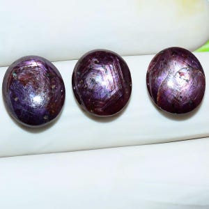 May include: Three round, cabochon-cut star rubies with a deep purple-red hue. Each gemstone displays a distinct six-rayed star effect across its polished, domed surface, with visible natural inclusions and internal crystalline patterns.