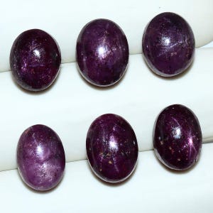 May include: Six oval-shaped, cabochon-cut rubies with a deep, dark purple-red hue. Each gemstone features a smooth, polished surface with visible natural inclusions and internal light reflections, arranged in two rows of three stones each.