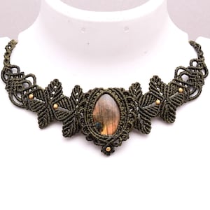 May include: A dark olive green macramé choker necklace featuring an oval-shaped labradorite gemstone at the centre. The intricate knotted cord design includes small metallic gold-toned beads integrated into the symmetrical patterns on both sides.