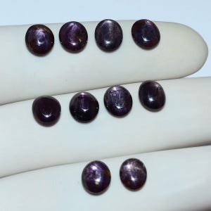 May include: A collection of ten oval-shaped, deep purple cabochon gemstones arranged in three rows. Each stone features a smooth, polished surface with a visible star-like light reflection, characteristic of star sapphires, displaying dark tones.