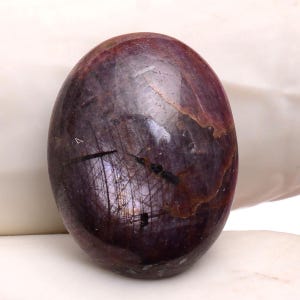 Op de afbeelding: Een gepolijste, ovaalvormige edelsteen met een diepe bordeauxrode kleur en subtiele interne patronen. De steen heeft een glad, reflecterend oppervlak met strepen van lichtere tinten en donkere insluitsels. De algehele uitstraling is rijk en elegant, wat wijst op een natuurlijk mineraal specimen.