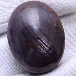 May include: A polished, oval-shaped gemstone with a deep purple hue, featuring subtle streaks and a glossy surface. The stone has a smooth, rounded form, and is set against a white background.