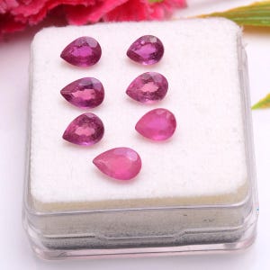 Puede incluir: Siete piedras preciosas rosas talladas en forma de pera, dispuestas sobre una base de espuma blanca dentro de una caja de presentación de plástico transparente. Cada piedra muestra un tono magenta vibrante con facetas internas visibles.