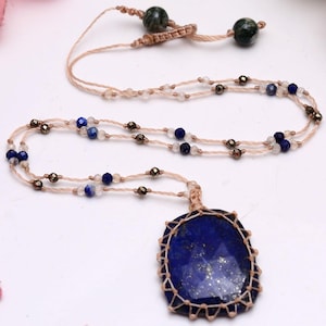 Könnte beinhalten: Eine Halskette mit einem großen, facettierten, tiefblauen Lapislazuli-Anhänger, der in eine hellbeige Makramee-Schnur eingefasst ist. Die Schnur ist mit kleinen, eingestreuten blauen Lapislazuli-Perlen, klaren Kristallen und metallisch dunkelgrauen Perlen verziert.
