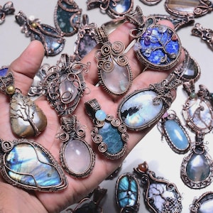 May include: A collection of handcrafted wire-wrapped pendants featuring various gemstones. The pendants showcase a range of colors, including blue, green, pink, and clear stones, set in intricate copper wire designs. The pendants are displayed in a hand.