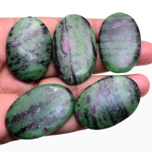 Puede incluir: Cinco cabujones ovalados pulidos hechos de zoisita con inclusiones de rubí. Cada piedra presenta una superficie lisa y brillante con un patrón moteado de verde vibrante, magenta profundo y vetas minerales negras oscuras en el material.