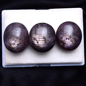 May include: Three oval-shaped, polished gemstones with a dark purple hue. The stones have a reflective surface with streaks of lighter color and some dark inclusions. They are displayed on a white surface.