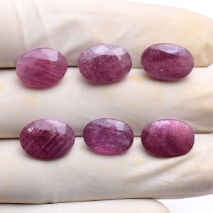 Puede incluir: Seis rubíes ovalados y facetados con un tono magenta rosado intenso. Cada piedra preciosa presenta una superficie pulida con inclusiones internas visibles y un corte de múltiples facetas que refleja la luz sobre la superficie.
