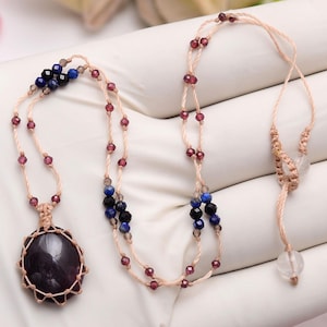 Op de afbeelding: Een ketting met een grote, donkerpaarse ovale steen als hanger in een lichtbeige geknoopt koord. Het koord is versierd met kleine, gefacetteerde rode, blauwe en zwarte kralen en bevat twee doorzichtige bolvormige kralen bij de sluiting.