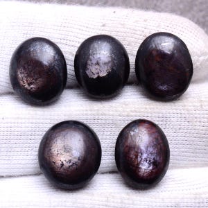 May include: Five dark, oval-shaped gemstones with a polished surface. The stones have a deep, almost black color with hints of reddish-brown and a subtle sheen. They are arranged on a white, textured background.