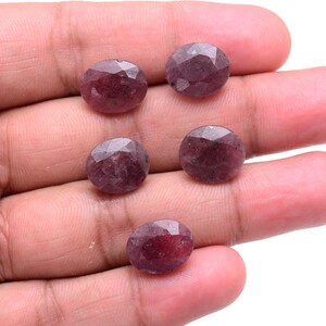 May include: Five oval-shaped, faceted gemstones with a deep reddish-purple hue. Each stone features a polished surface with multiple geometric facets that reflect light, showcasing a translucent quality and a consistent, dark berry-toned colour.