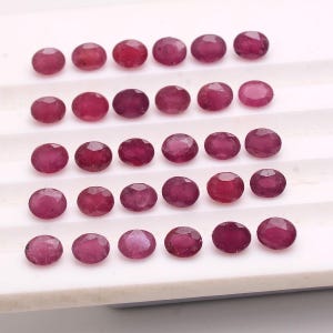 May include: A collection of twenty-nine oval-shaped, faceted rubies arranged in five horizontal rows. Each gemstone displays a deep, saturated magenta-red hue with a polished, reflective surface, showcasing multiple geometric facets.