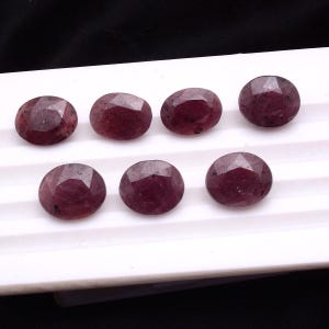 Puede incluir: Siete gemas ovaladas y facetadas en tonos rojo oscuro y burdeos están dispuestas sobre una superficie blanca. Las gemas tienen una superficie pulida y reflectante, y probablemente sean rubíes, adecuadas para la fabricación de joyas.