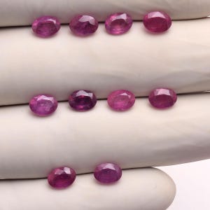 May include: Ten oval-cut, faceted rubies in a vibrant magenta-pink hue. The gemstones are arranged in three rows, displaying a polished, reflective surface with clear internal facets. Each ruby exhibits a consistent oval shape and deep color.