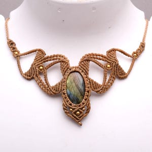 May include: A macrame necklace featuring a central oval labradorite gemstone with iridescent blue and green flashes. The stone is encased in intricate tan cord knotting with symmetrical geometric patterns and small embedded brass beads.