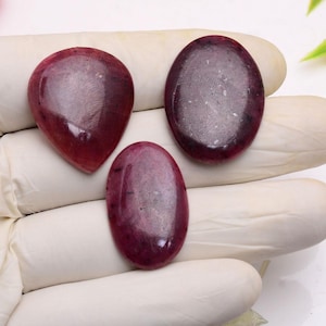 May include: Three polished, oval-shaped gemstones in varying sizes. The stones are a deep, rich burgundy color with subtle variations in tone. One is teardrop-shaped. The gemstones are displayed on a white surface.