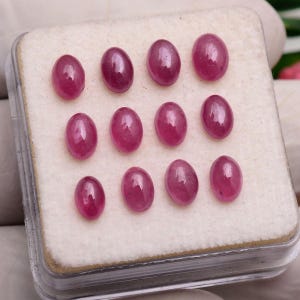 May include: A collection of twelve oval-shaped, cabochon-cut rubies with a deep pinkish-red hue and a smooth, polished surface. The gemstones are arranged in a grid pattern on a white foam insert inside a clear, square plastic display case.