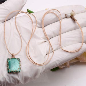 May include: A square, green malachite pendant necklace with a woven gold-colored wire frame. The pendant hangs from a tan cord. A second necklace with two small, round, off-white beads is also visible.